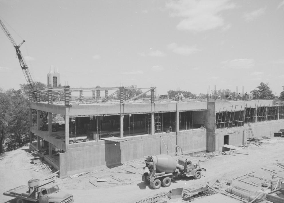 Black-and-white photograph of library walls being built. The facade is not yet present. A crane rises above the trees, and a cement truck drives by.