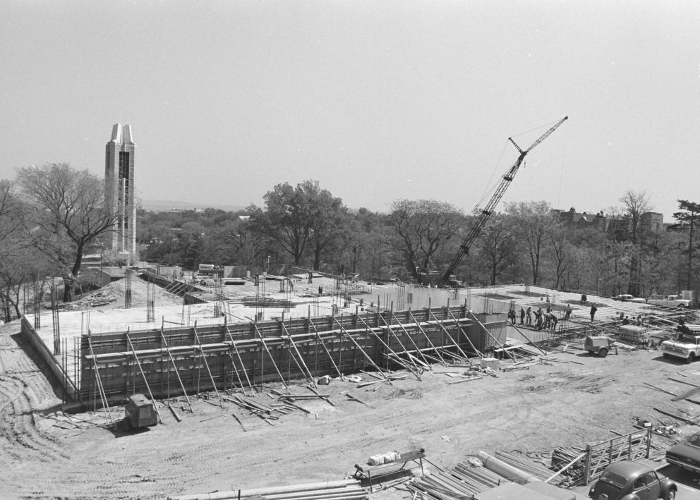 Black-and-white photograph of the library foundation and a partial wall. A crane rises above the trees and the Campanile is prominent in the background.