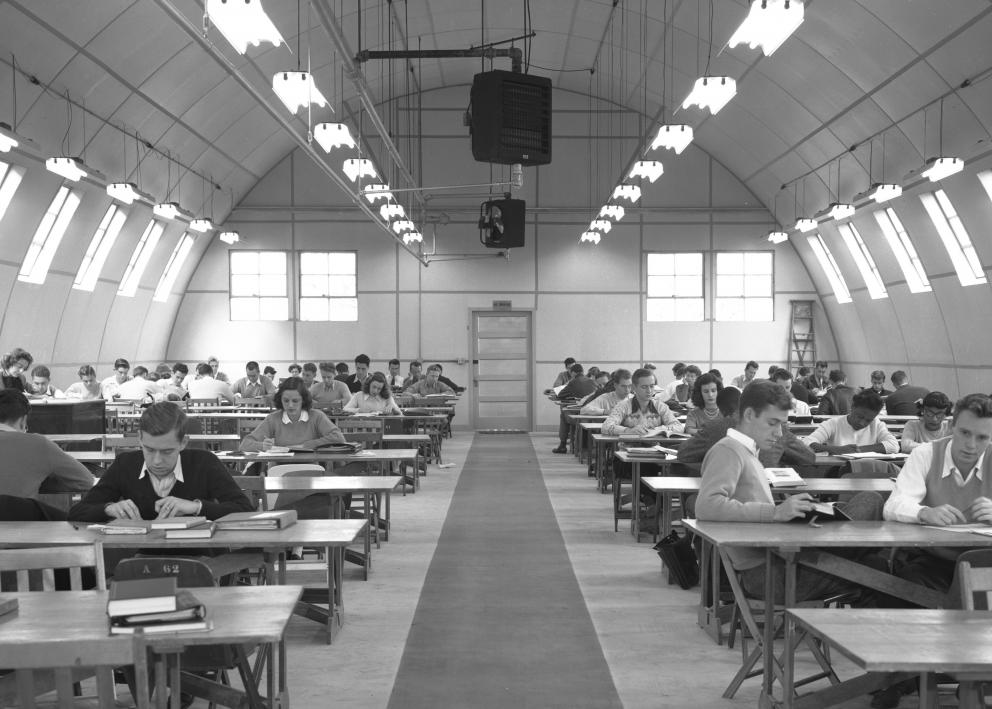 Black-and-white photograph of students sitting at long tables on either side of a middle walkway. Lights hang down from the domed ceiling.