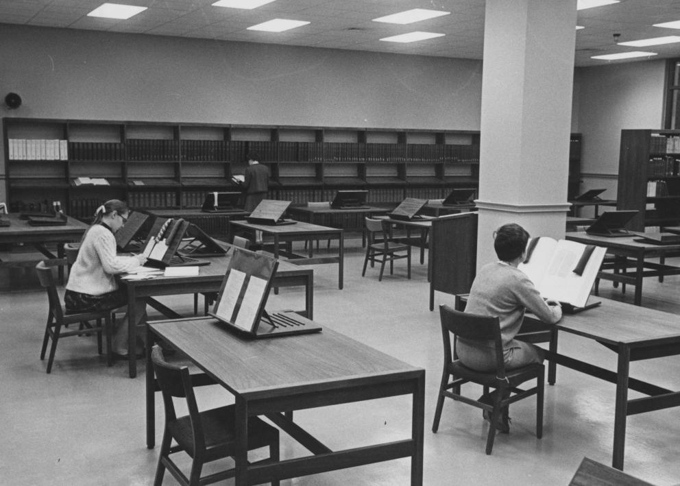 Black-and-white photograph of tables, chairs, and bookcases in the old Reading Room. Two researchers sit at tables viewing books propped up and open on stands.