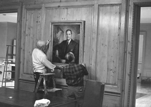 Black-and-white photograph of two men with their backs to the camera, standing behind a desk hanging a painting – a short ladder between them.