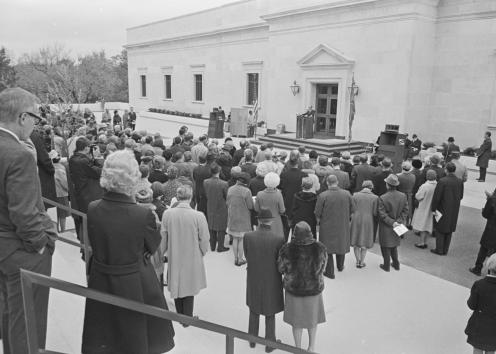 Black-and-white photograph of people standing on the library terrace facing a speaker at a podium on the library's front steps.