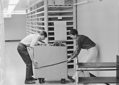 Black-and-white photograph of two people rolling a cart of books. Empty shelves are visible behind them.
